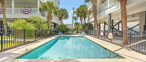 A swimming pool with lounge chairs, palm trees, and a building with American flags.