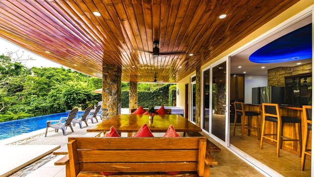 Uma área de refeições ao ar livre com piscina, móveis de madeira e vista para o verde.
