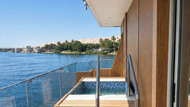 Una terraza con barandilla de vidrio que da a una masa de agua, una cubierta de madera y una puerta de cristal que conduce a un espacio interior.