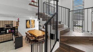 A modern interior with a wooden dining table, a staircase with black railings, and a kitchen area.
