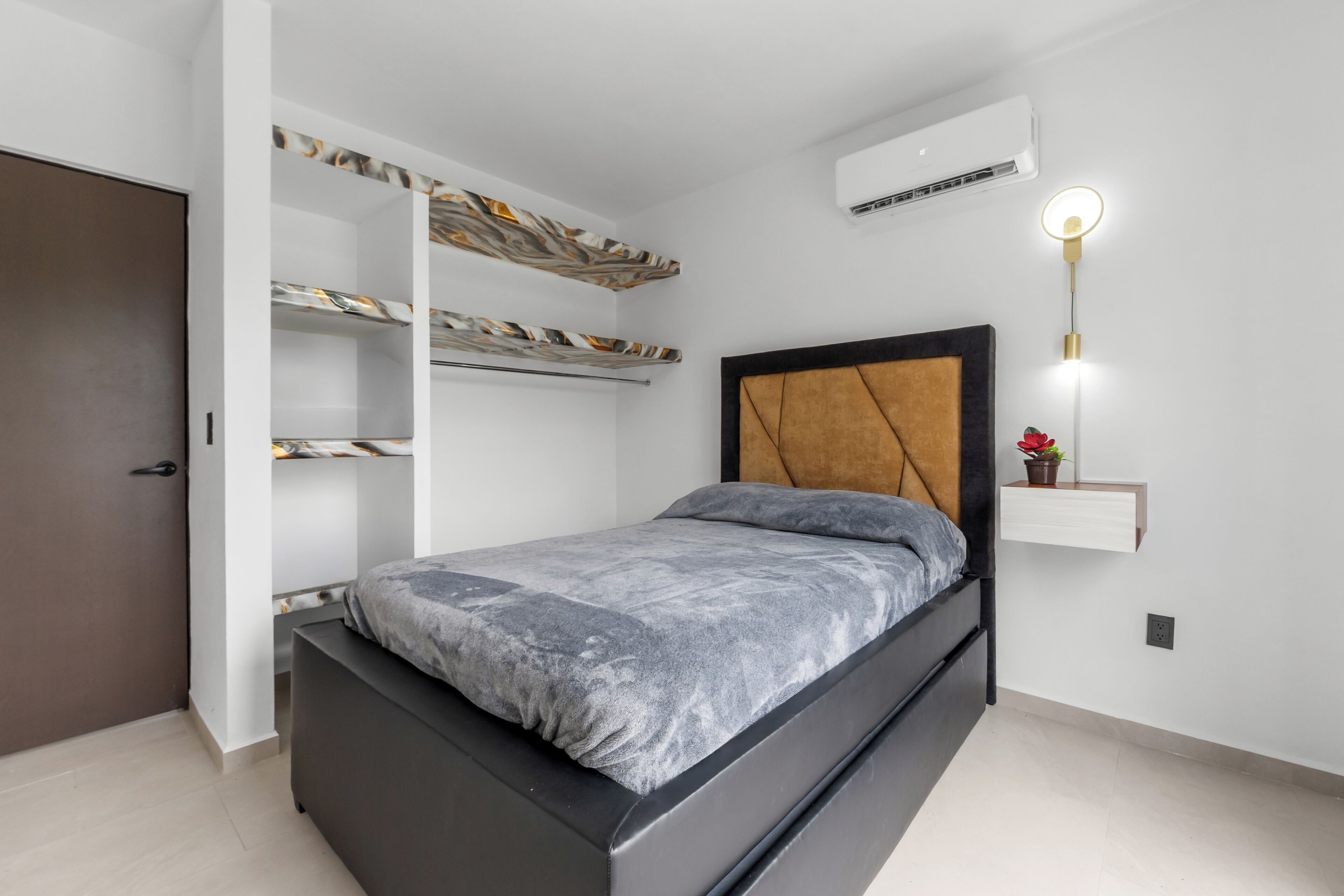 A bedroom with a bed, built-in shelving, an air conditioning unit, and a wall-mounted light fixture.
