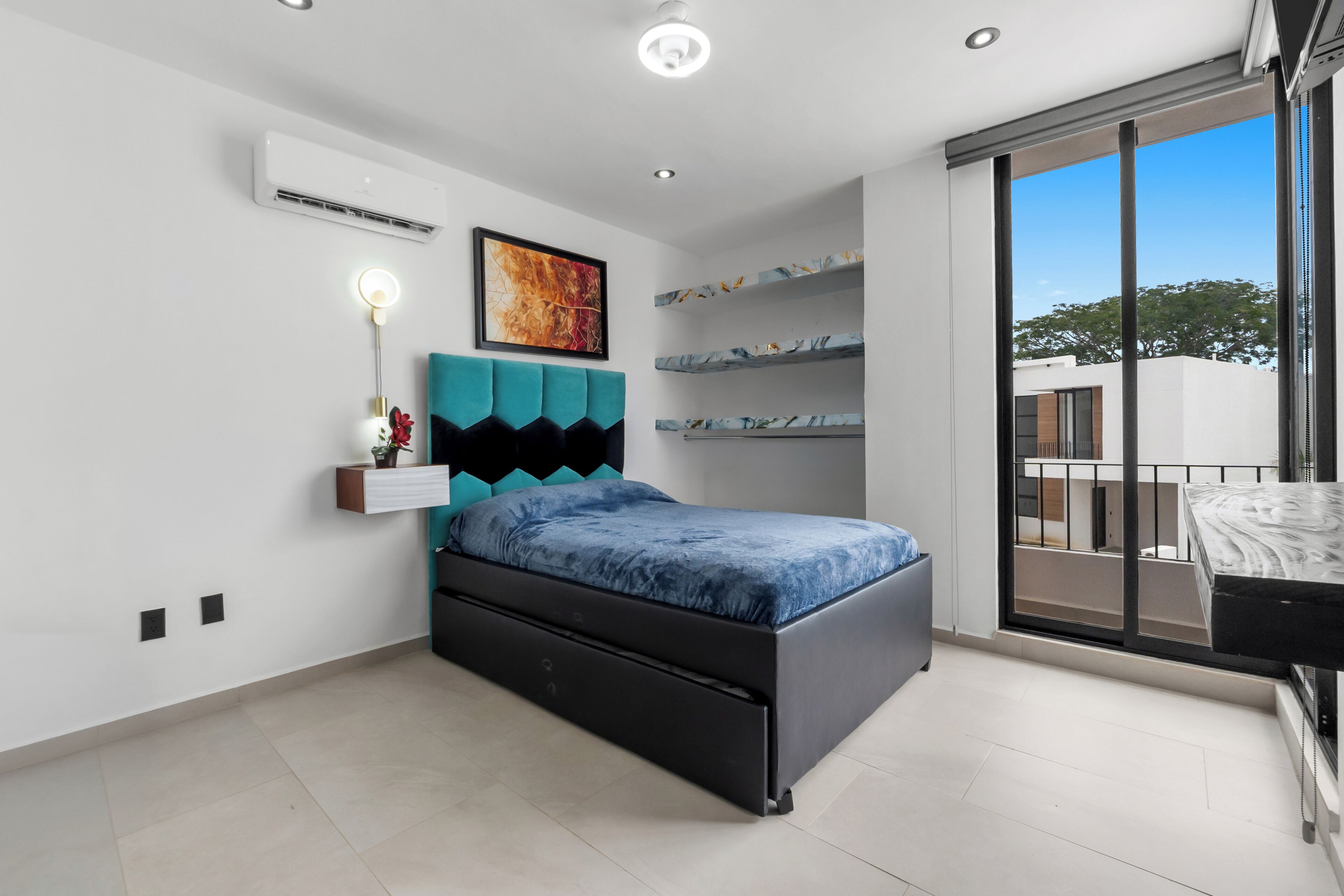 A modern bedroom with a bed, a wall-mounted air conditioner, a ceiling light, and a large window with a view of the outside.