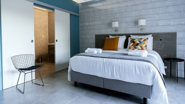 Quarto de hotel moderno com uma cama grande, uma cadeira e um banheiro visível por uma porta aberta.
