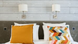 Cama com roupa de cama branca, duas luminárias fixadas na parede e cabeceira de madeira.