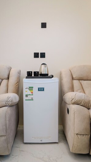 A compact refrigerator with a control panel and a kettle on top, placed between two beige armchairs in a room with a plain wall and two electrical outlets. - 2BR Serviced Apartment in Al Rimal  Near PNU & Riyadh Airport (الرياض)