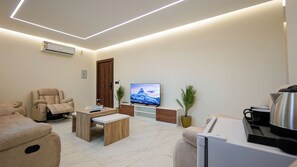 Ein modernes Wohnzimmer mit einer Couch, einem Relaxsessel, einem Couchtisch, einem Fernseher und einer kleinen Küchenzeile mit Wasserkocher.