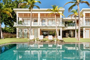 Une maison moderne à deux étages, dotée d’une piscine, de palmiers et d’un espace salon extérieur.