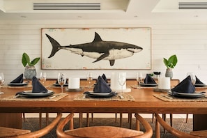 Mesa de comedor preparada para seis personas, con individuales, platos y vasos, un cuadro grande de un tiburón en la pared y dos plantas en macetas.