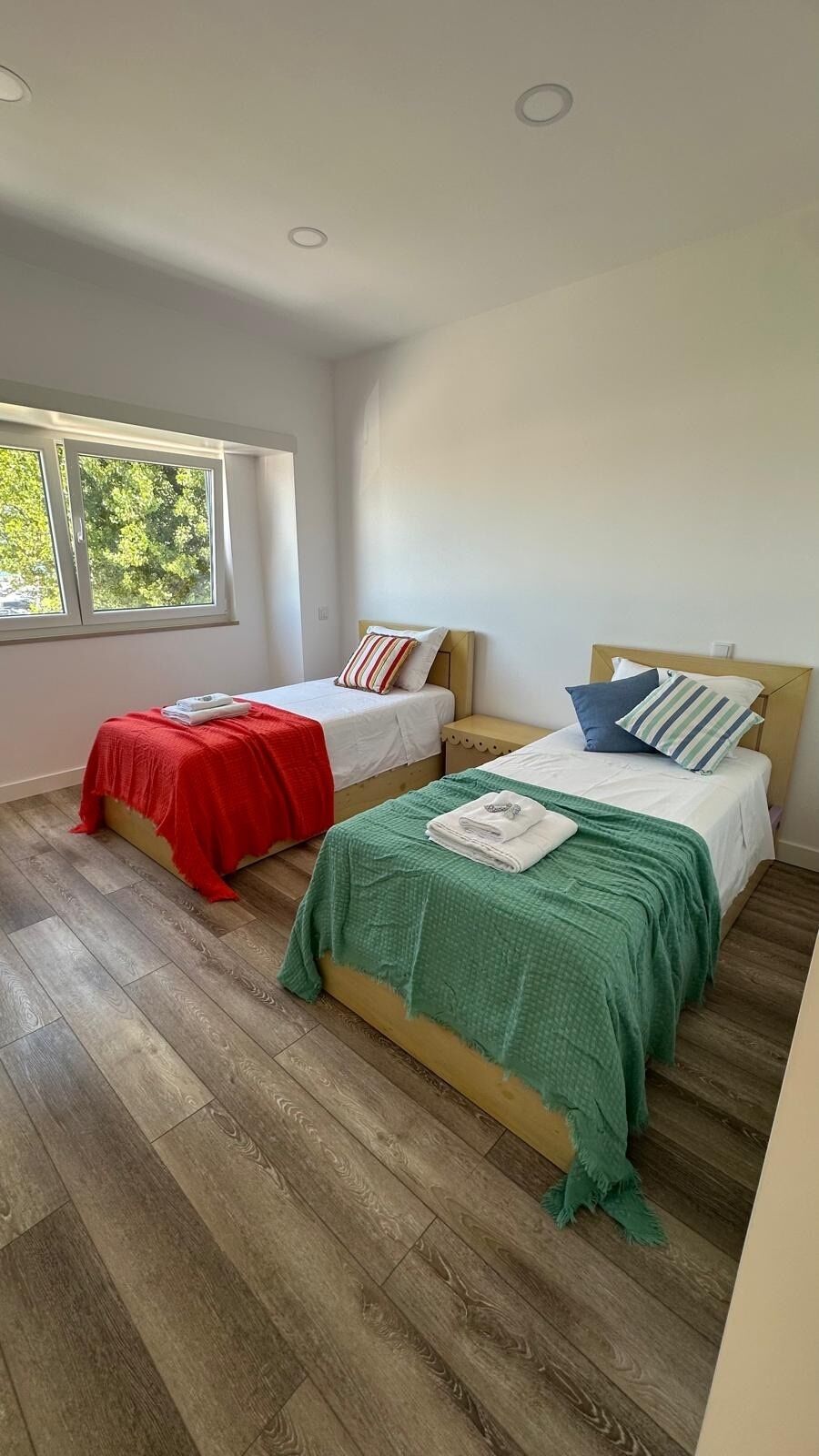 A room with two beds, wooden flooring, a window with a view of trees, and a ceiling with recessed lighting.