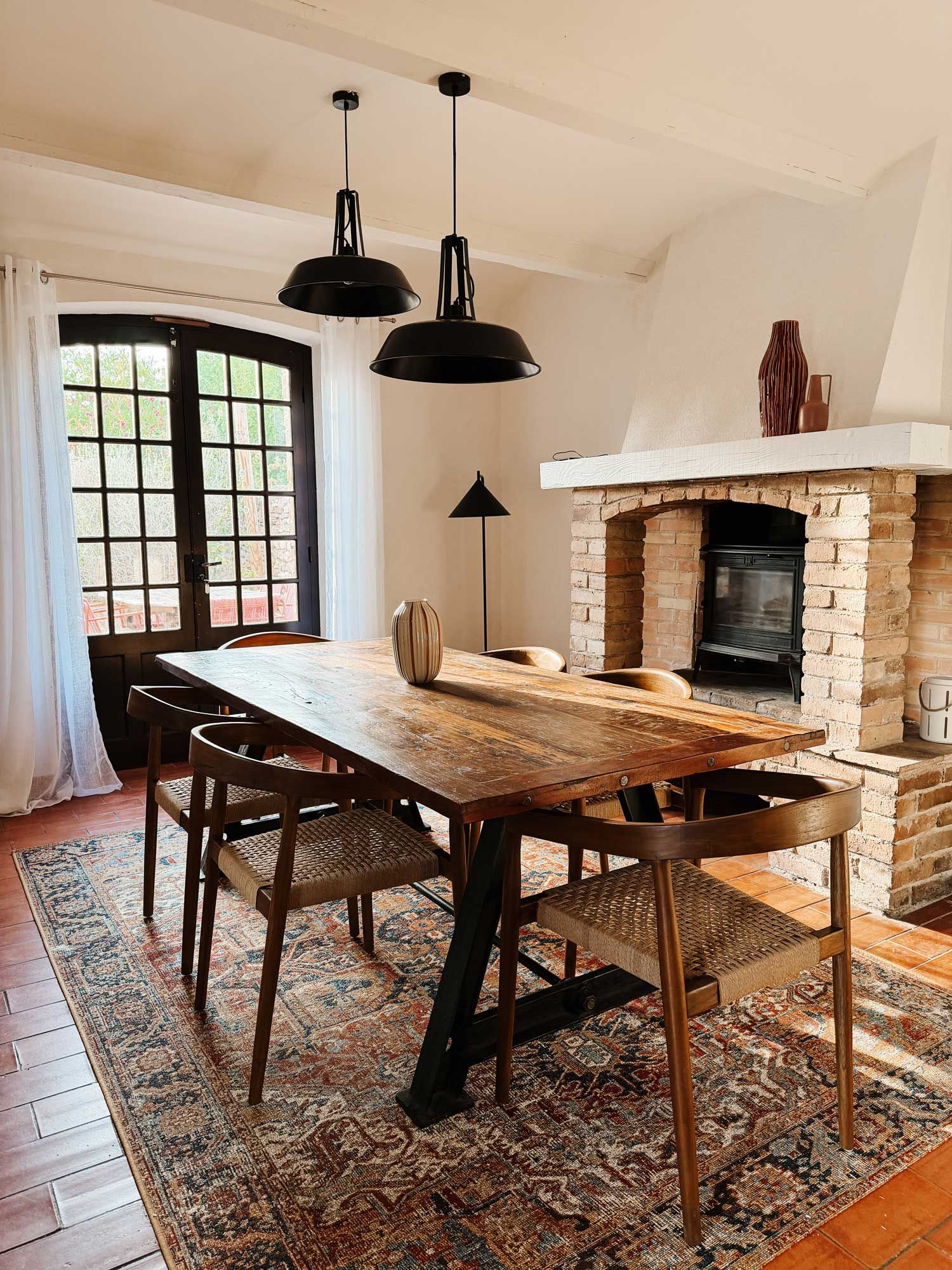 Un comedor con mesa de madera, sillas, chimenea y lámparas colgantes.