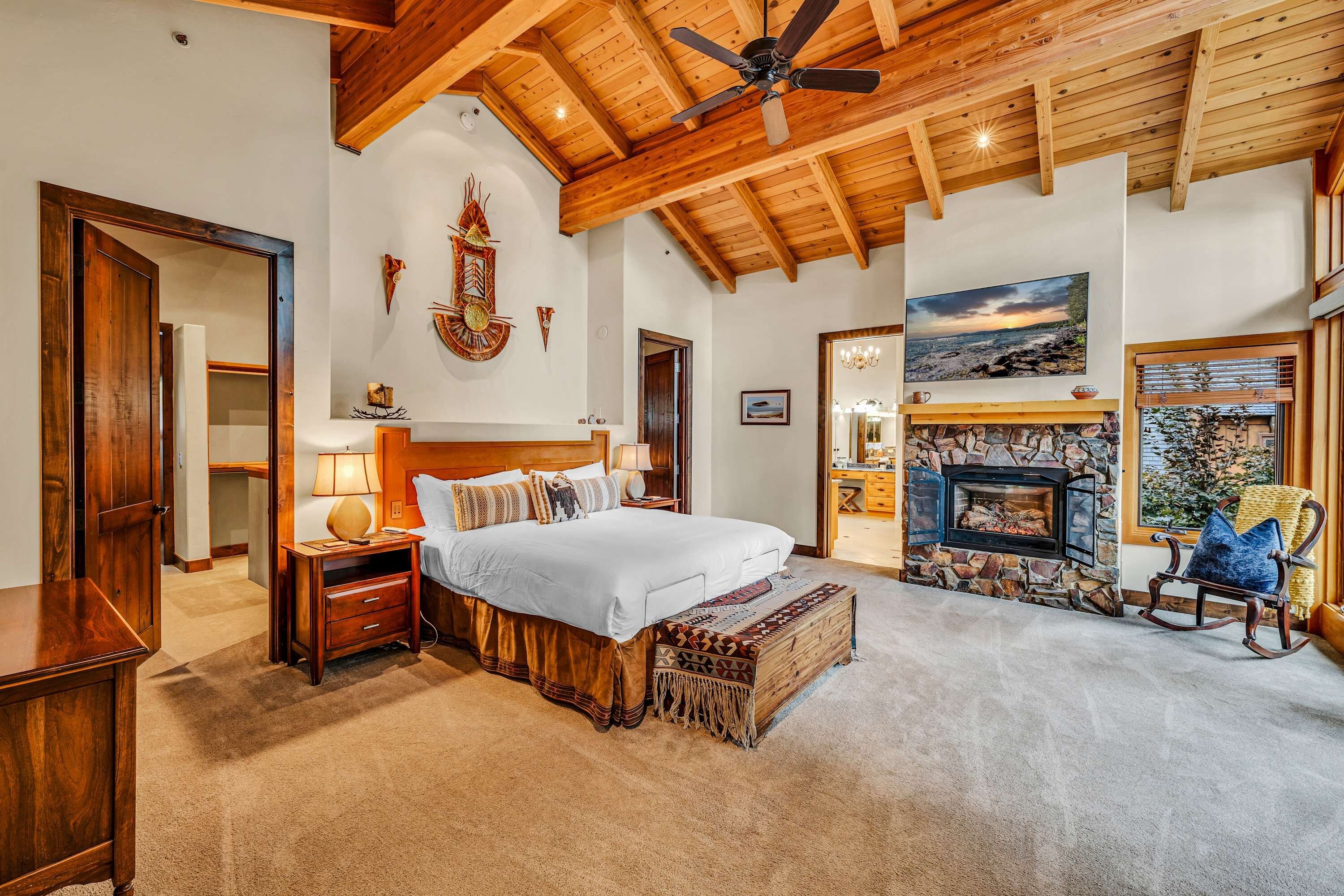 A bedroom with a large bed, a stone fireplace, a ceiling fan, and a view of the outdoors through a window.