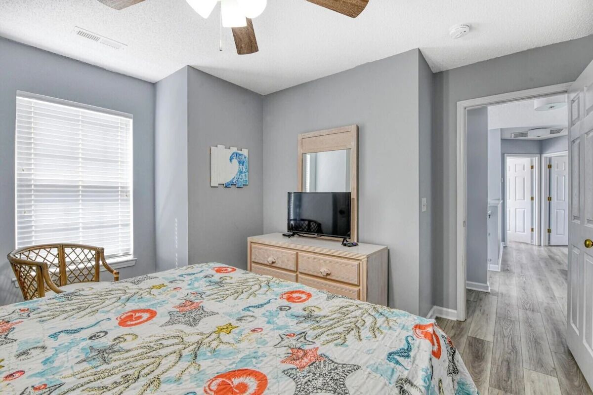Een slaapkamer met een bed, een dressoir en een spiegel. Er is een raam met zonwering en een deur naar een andere kamer.