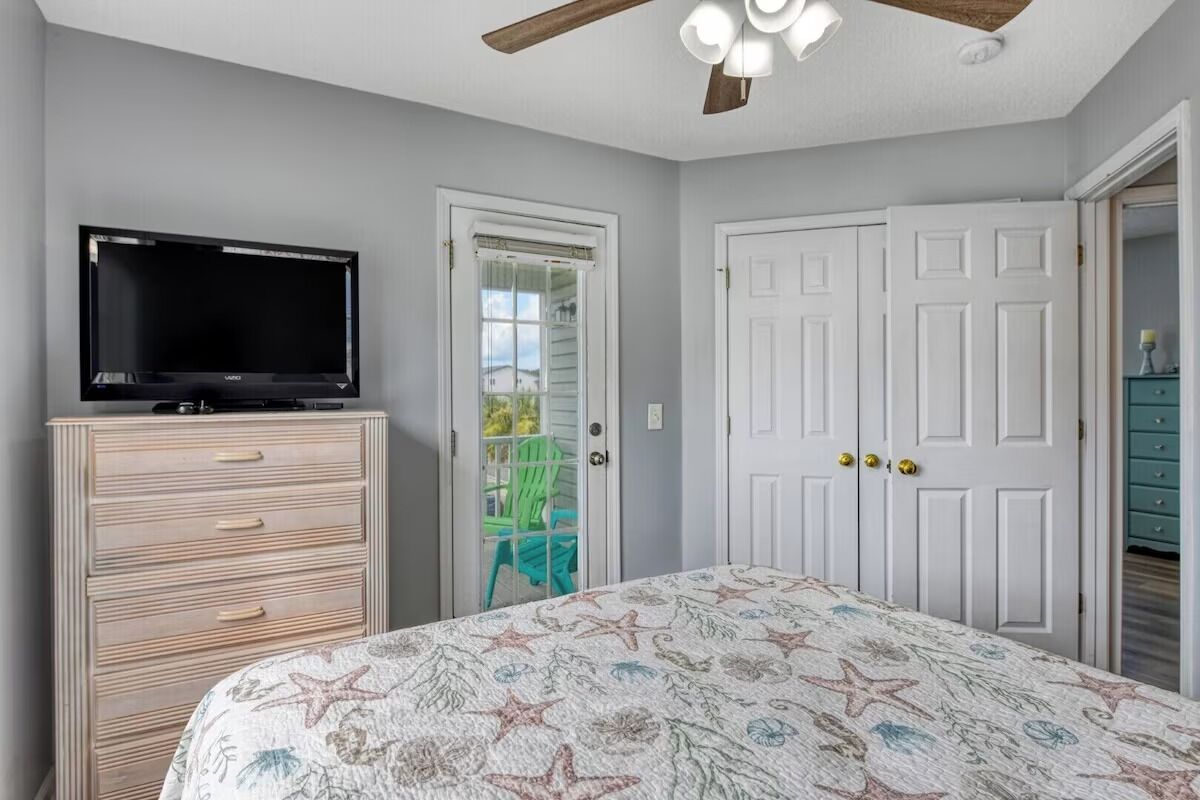 A bedroom with a bed, a television on a dresser, a ceiling fan, and a door leading to a balcony.