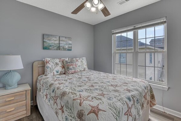 A bedroom with a bed, a nightstand, a lamp, a ceiling fan, a window, and a wall-mounted artwork.