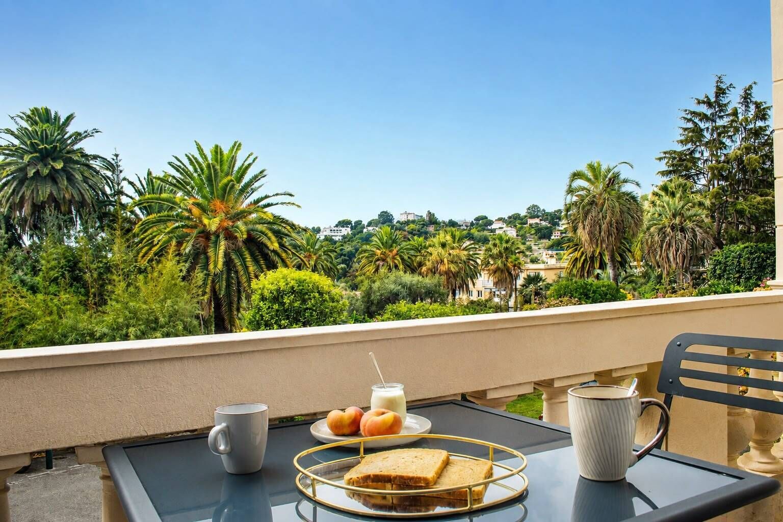 Un balcon avec une table préparée pour le petit-déjeuner, comprenant des toasts, des fruits et une boisson, donnant sur un jardin luxuriant avec des palmiers et un ciel dégagé.