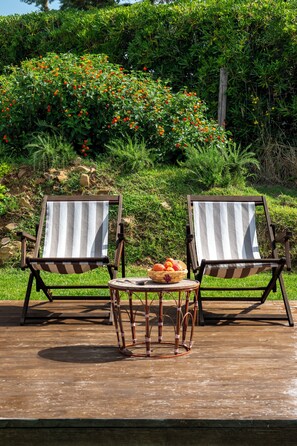 Twee gestreepte lounge fauteuils met een klein tafeltje en een mand vol fruit ertussen, op een houten terras met een tuin en bloeiende planten op de achtergrond.