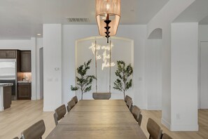 Uma sala de jantar moderna com uma mesa de madeira comprida, duas plantas em vasos e um letreiro de neon na parede.