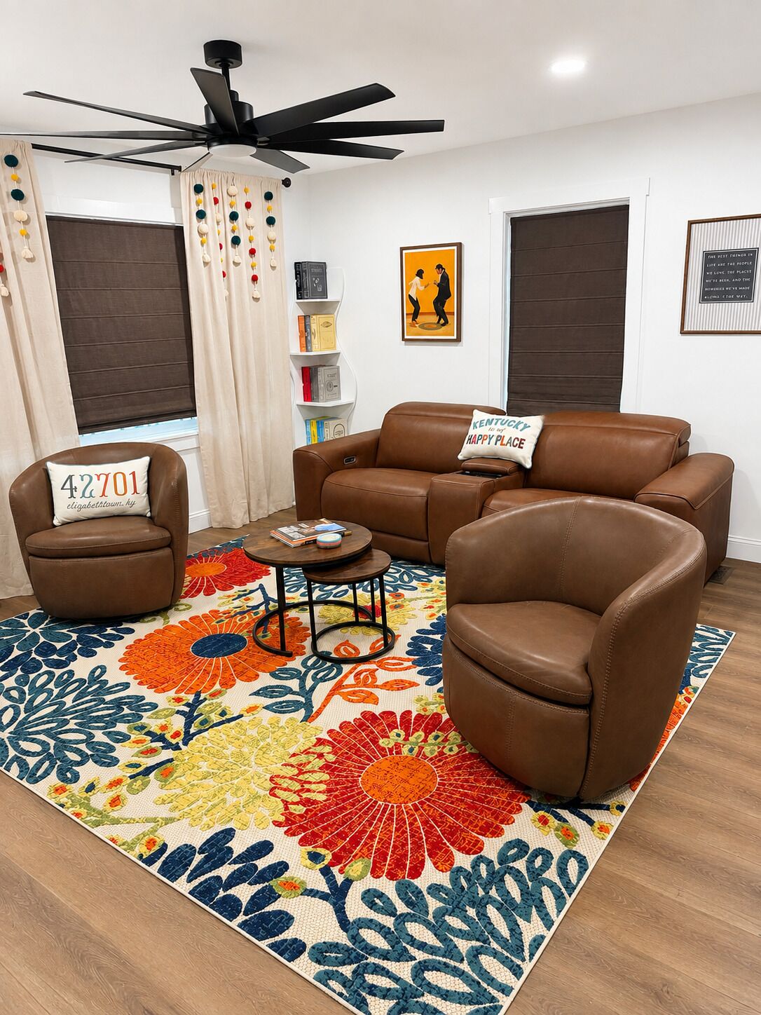 Ein Wohnzimmer mit einem braunen Ledersofa, zwei braunen Ledersesseln, einem Couchtisch und einem farbenfrohen Teppich mit Blumenmuster. Es gibt einen Deckenventilator, ein Bücherregal und ein gerahmtes Bild an der Wand.