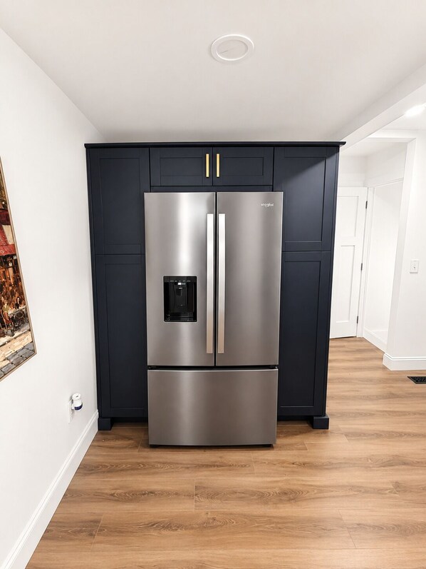 A stainless steel refrigerator with a water dispenser, set against dark cabinetry in a kitchen with wooden flooring.
