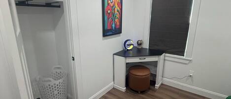 Ein Zimmer mit Deckenventilator, Fenster mit Jalousien, Schreibtisch mit Stuhl, ein farbiges Gemälde an der Wand und ein weißer Kleiderschrank mit Kleiderstange.