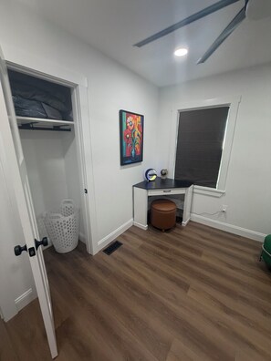 A room with a ceiling fan, a window with blinds, a desk with a chair, a colorful painting on the wall, and a white closet with a hanging rod.