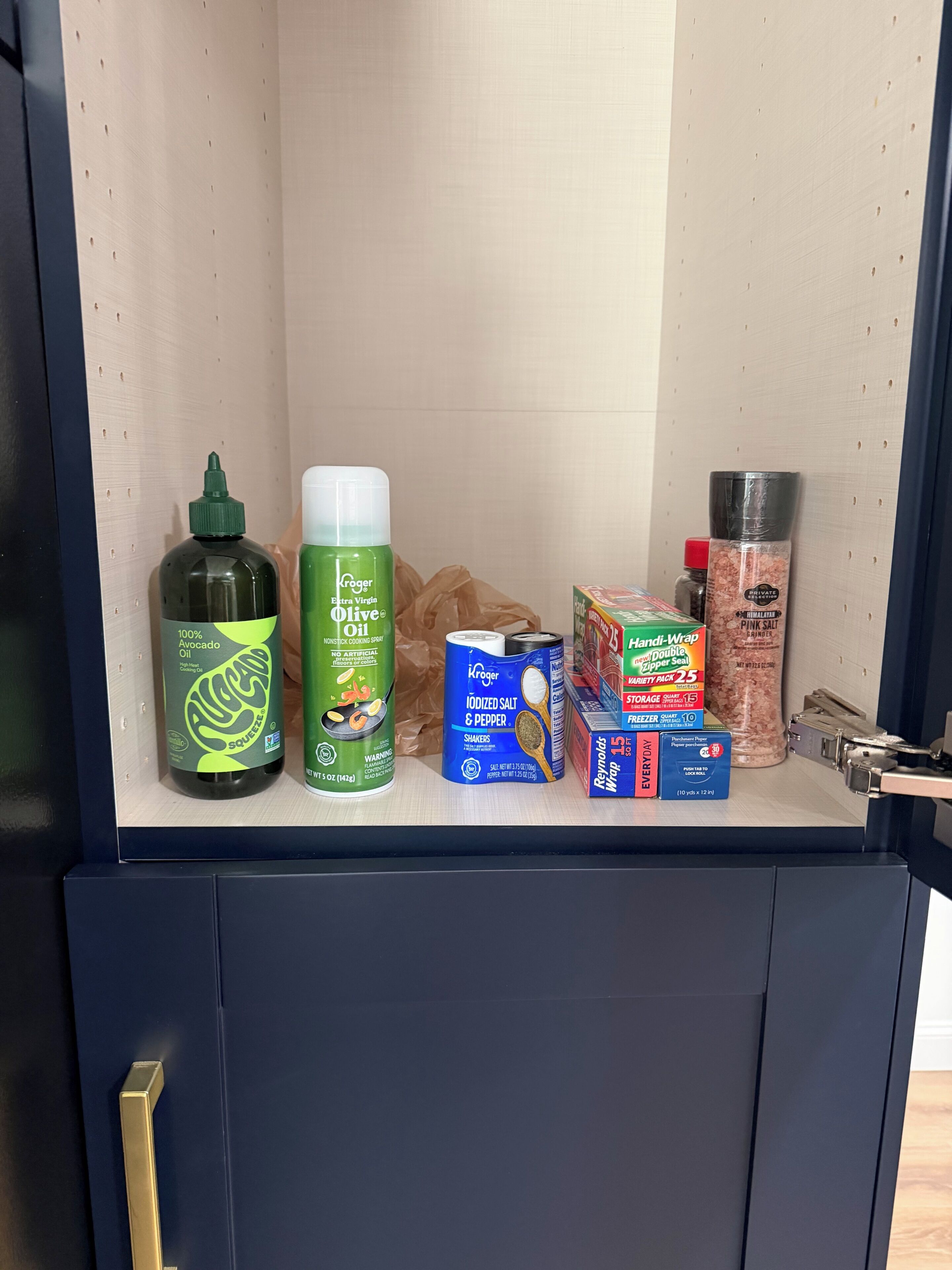 Un pensile da cucina con diversi oggetti, tra cui una bottiglia di olio antiaderente, una lattina di sale iodato, una scatola di Reynolds Wrap e un contenitore di sale rosa dell'Himalaya.