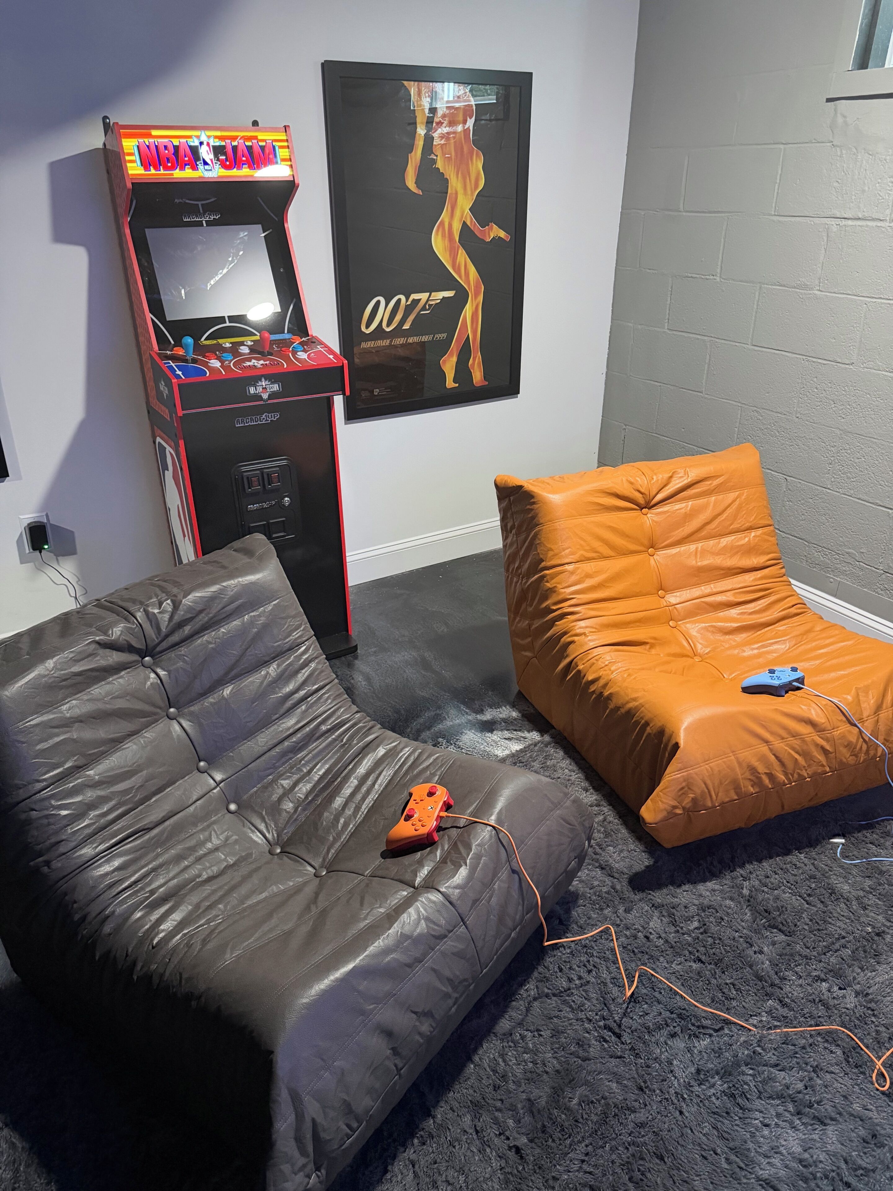 Ein Zimmer mit Spielautomaten, einem Poster, einem grauen Sofa und einem orangefarbenen Ottomanen.