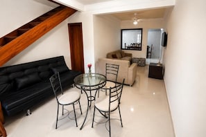 A living room with a black sofa, a glass table with chairs, a wooden staircase, and a view into another room with a sofa and a TV.