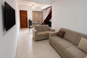 A living room with a flat-screen TV mounted on the wall, a sofa, a dining table with chairs, and a staircase. - Charming Chalet in the Penedo Mountains (Itatiaia)