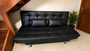 A black leather sofa with a tufted backrest and four wheels, positioned in a room with a wooden staircase and a potted plant.