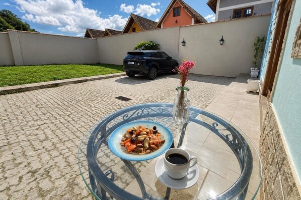 A glass table with a plate of food and a cup of coffee, a car parked in a courtyard, and a building with a tiled roof in the background. - Charming Chalet in the Penedo Mountains (Itatiaia)