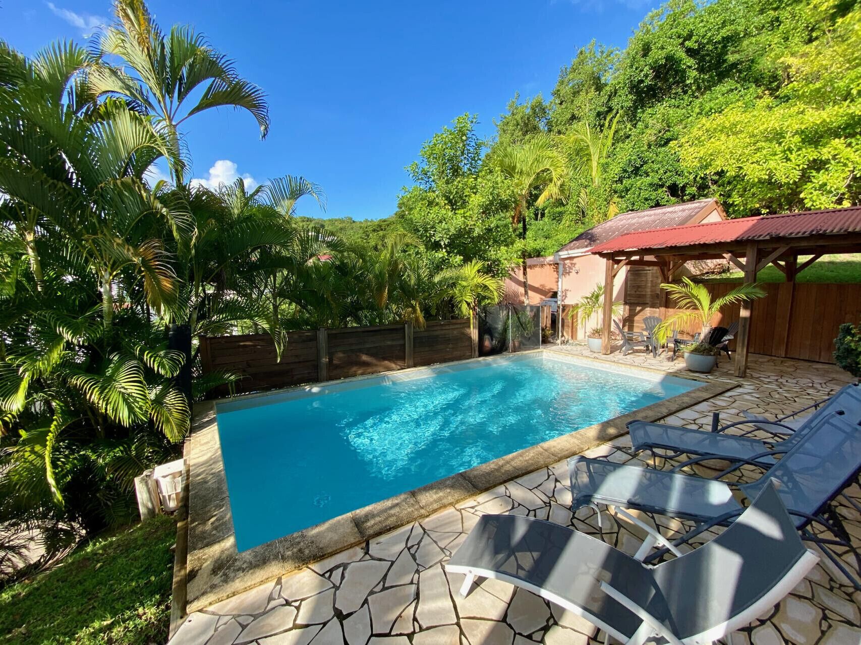 Piscina com deck de pedra, rodeada por vegetação exuberante e estrutura de madeira.