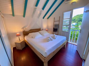 A bedroom with a wooden bed, white bedding, a bedside lamp, and a view of greenery through a window. - Sea and pool view, fully equipped villa for 8 (Les Trois-Ilets)