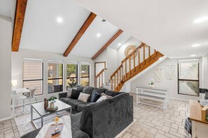 A spacious living area with a grey sofa, a coffee table, and a staircase with wooden railings. - Charming 2BR Home | Fireplace & Outdoor Lounge (Abilene)