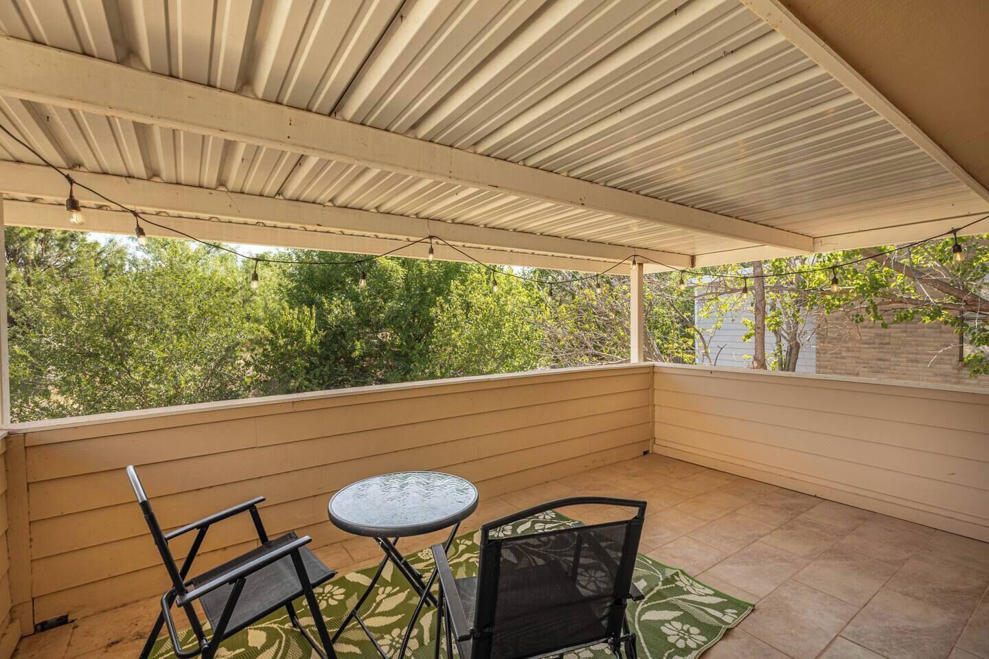 A covered patio with a table and chairs, a view of trees, and a brick wall in the background.