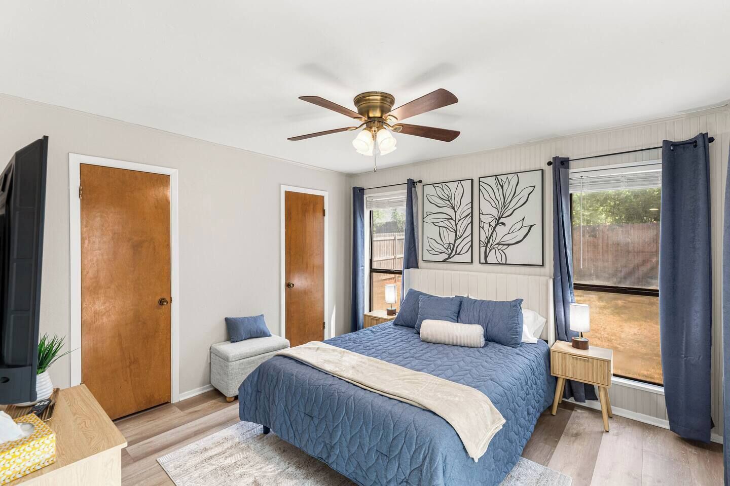 A bedroom with a bed, a ceiling fan, a nightstand, a bedside table, a TV, and a window with curtains.