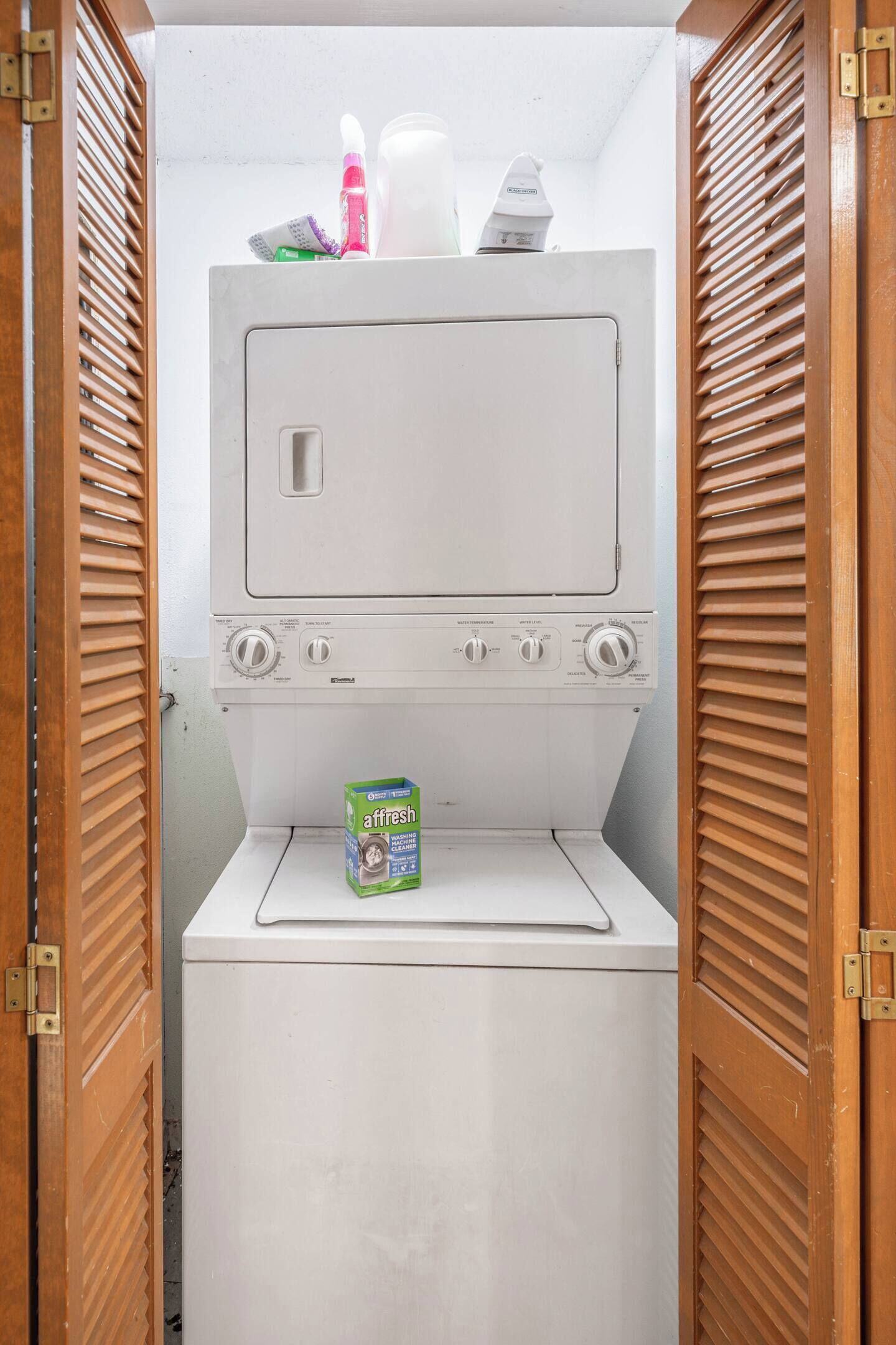 A compact washing machine with a detergent box labeled 'affresh' in a small laundry room with wooden shutters.
