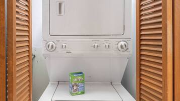 A compact washing machine with a detergent box labeled 'affresh' in a small laundry room with wooden shutters.
