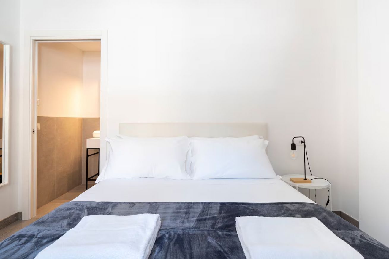 Una camera d'albergo con un letto, un comodino con una lampada e una porta che dà su un'altra stanza.