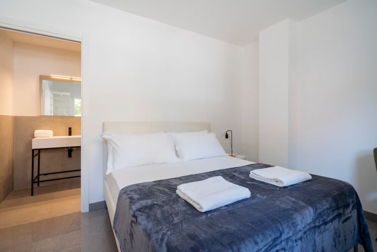 Una camera d'albergo con un letto, un comodino, uno specchio e un bagno con lavandino e asciugamano.