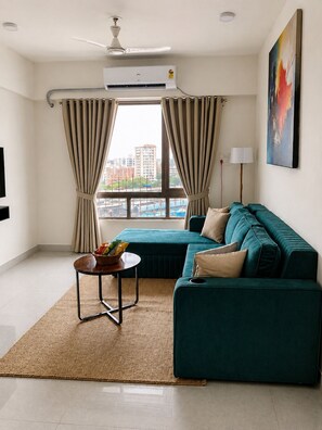 A modern living room with a teal sofa, a small round table with a basket, beige curtains, and a view of buildings through the window. - Premium 2-bedroom apartment in the hearts of Mumbai! (Mumbai)