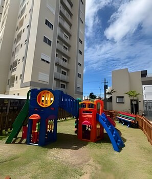 A playground with slides and climbing structures, surrounded by a grassy area and tall residential buildings. - Apto 2 Qtos I Suíte I Lazer completo (Cabedelo)