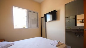 Ein Hotelzimmer mit Bett, Wandfernseher, Fenster mit Jalousien sowie Badezimmer mit Spiegel und Waschbecken.