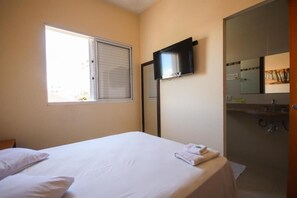 A hotel room with a bed, a television mounted on the wall, a window with blinds, and a bathroom with a mirror and sink. - Hotel Paulista (São José do Rio Pardo)