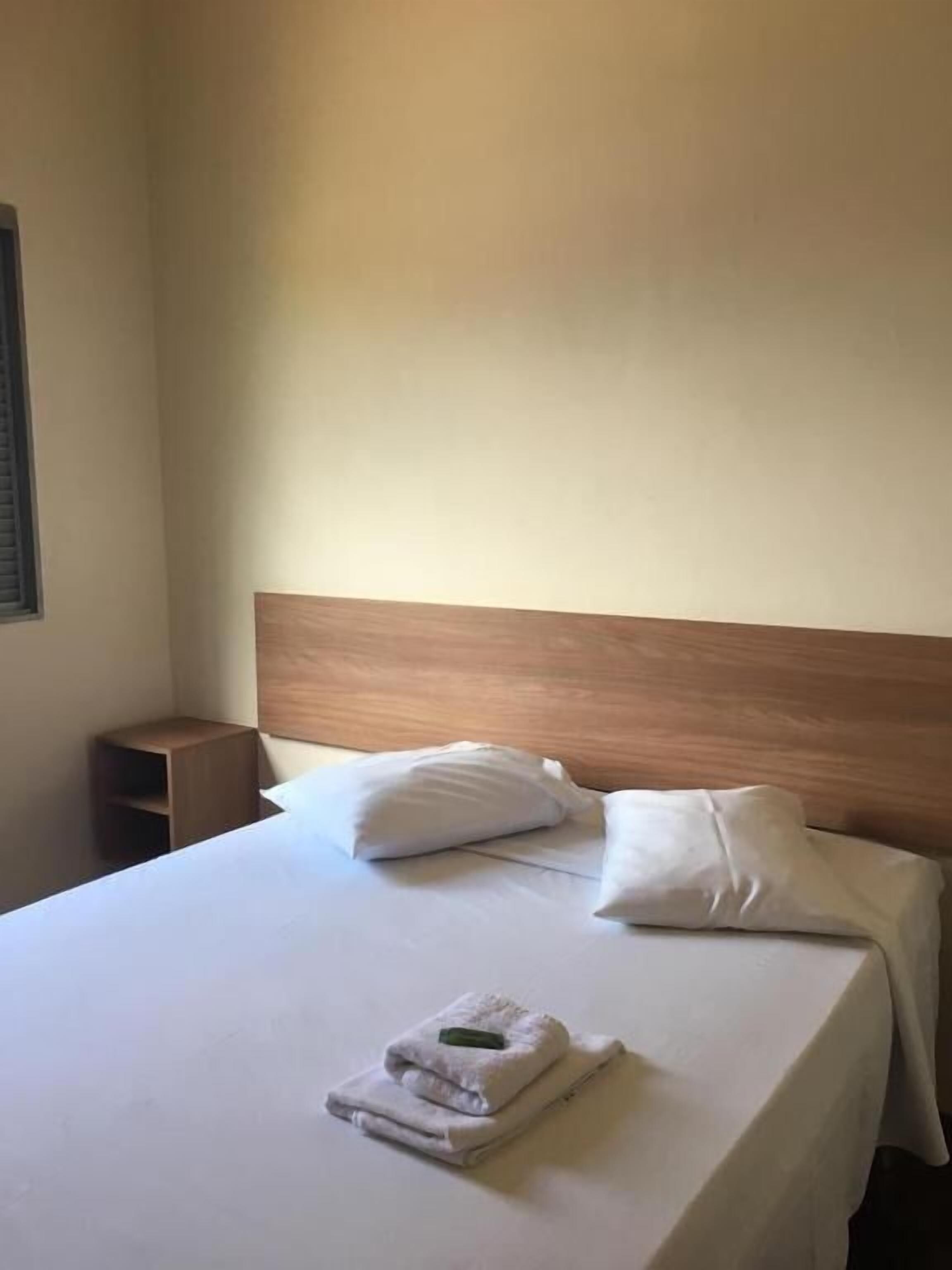 Quarto de hotel com uma cama bem arrumada, cabeceira de madeira e uma pequena mesa de cabeceira.