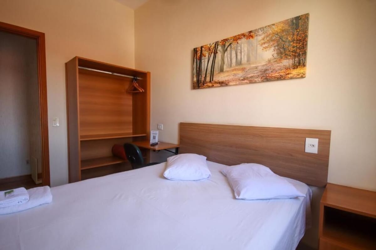 A hotel room with a bed, a painting of a forest, a wooden wardrobe, and a desk.
