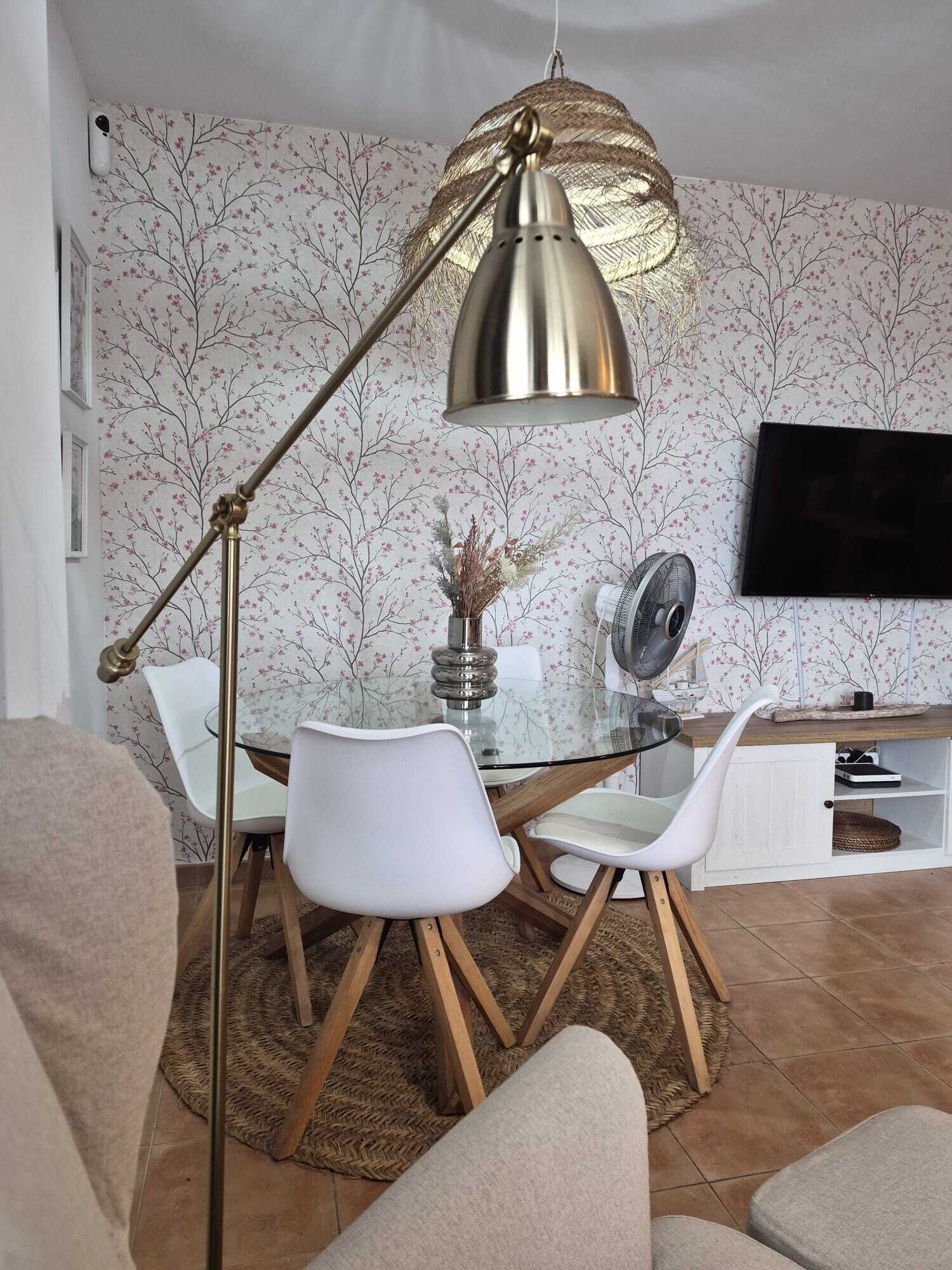 A modern living room with a glass dining table, white chairs, a floor lamp, and a flat-screen TV mounted on the wall.