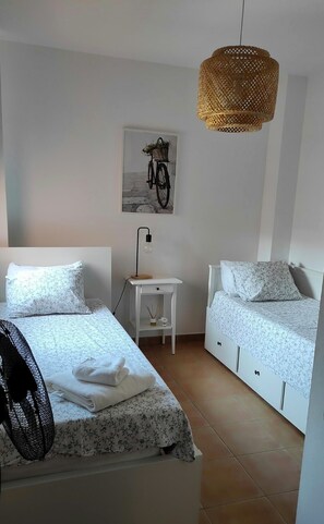 Ein Schlafzimmer mit zwei Betten, einem Nachttisch mit Lampe, einer Hängeleuchte aus Weidwerk und einem an der Wand hängenden Bild von einem Fahrrad.