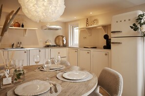 A dining area with a round table set for four, a kitchen in the background, and a refrigerator with the brand name visible. - 4P Modern Chalet with Private Sauna - Fenced Garden in a Wooded Area - BBQ (Putten)