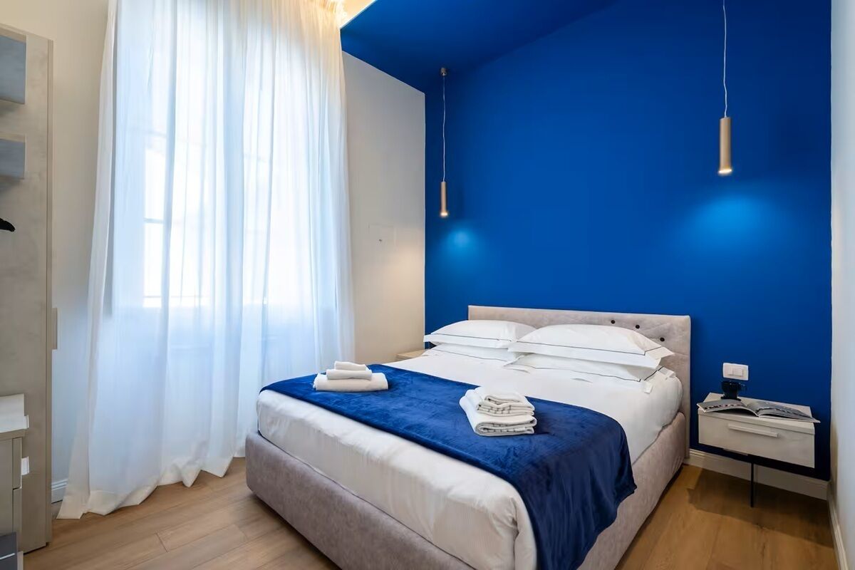 Een slaapkamer met een bed, een nachtkastje, een raam met gordijnen en een blauwe muur.